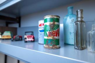 Collection of historic glass bottles, beverage cans, and toy vehicles displayed on a museum shelf.